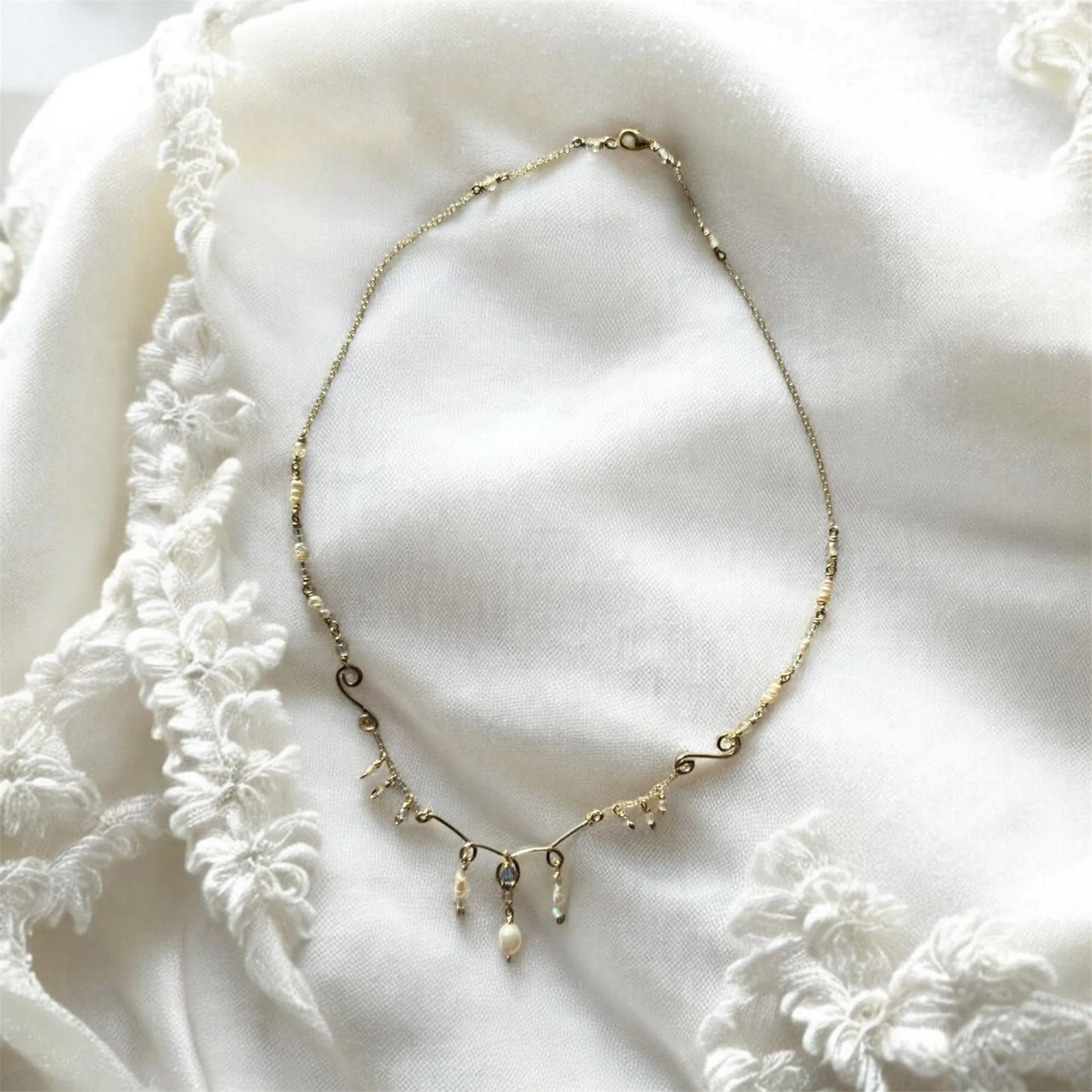 A delicate gold chain necklace with seed pearls and small dangling pearl drops, laid on cream lace.