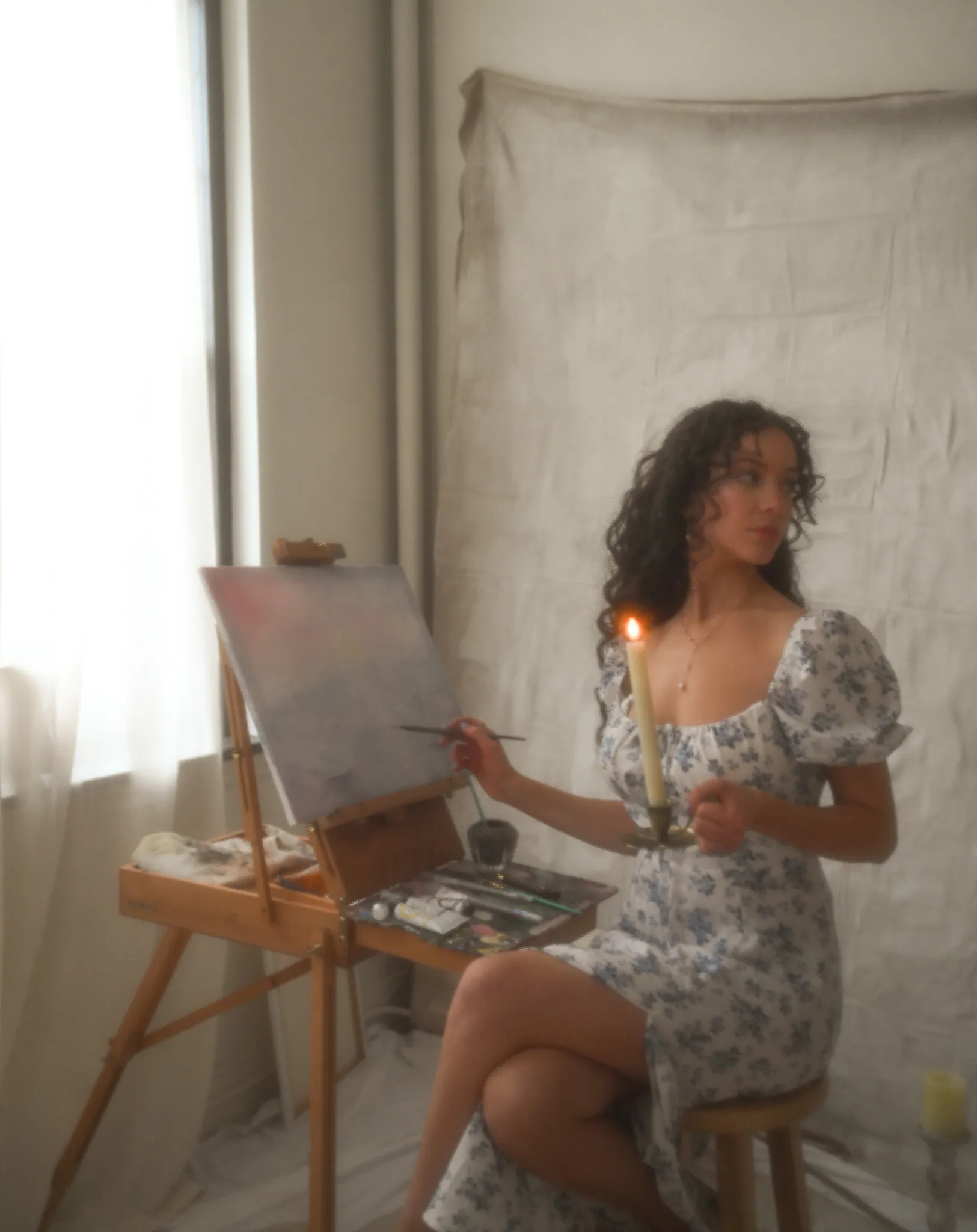 Abigail seated at her easel, holding a candle beside an oil painting.
