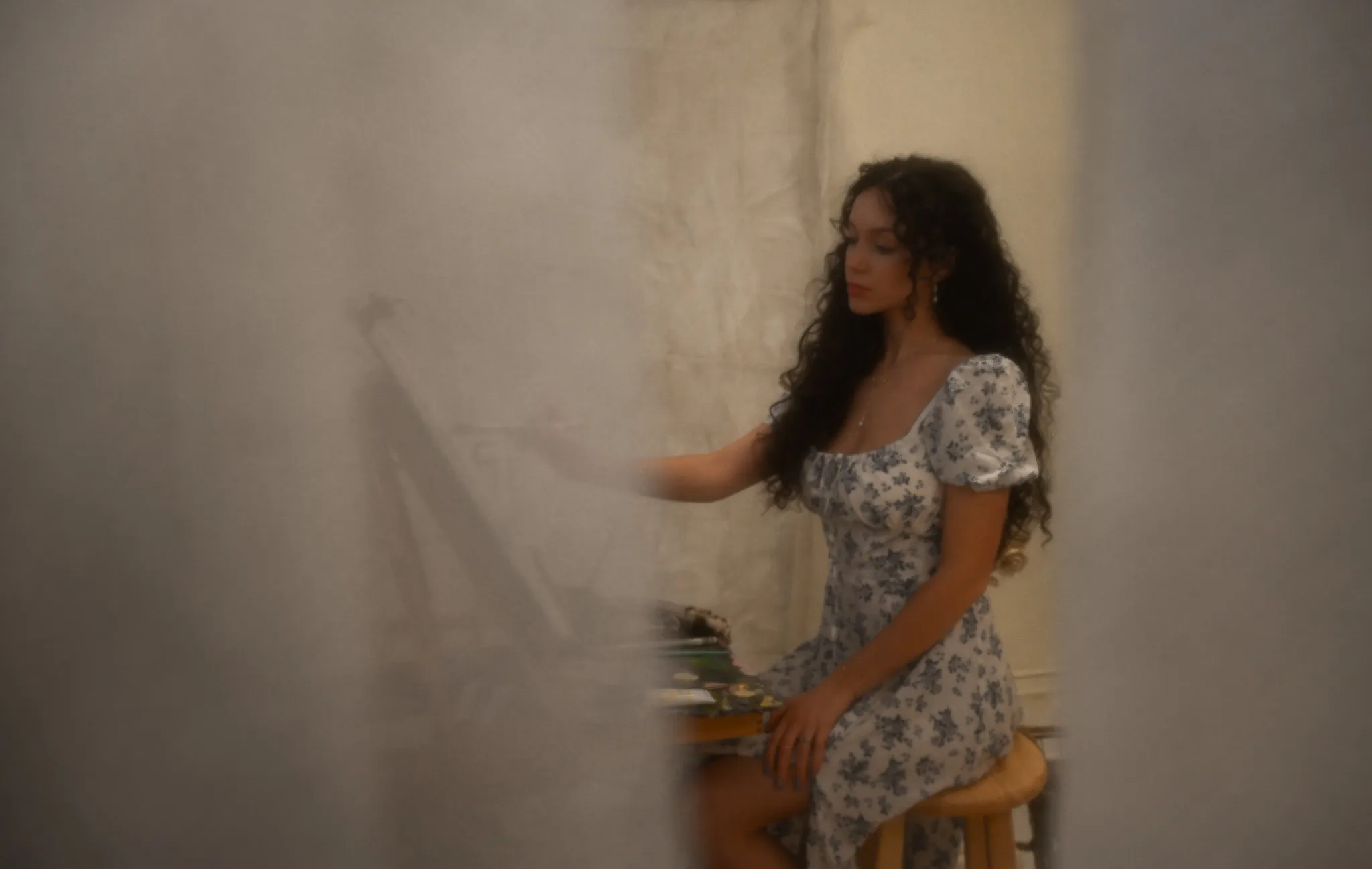 The artist seated at her easel on a wooden stool, in soft focus.